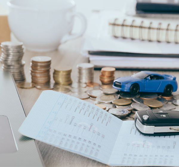 A close-up of A stock of coins, a miniature blue car, a car key, and a receipt on top of a table against a blurry cap and stock of books in the background.